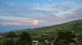 Pemandangan rumah adat panggung beratap alang-alang di Desa Adat Praijing, Pulau Sumba, dengan latar belakang perbukitan hijau saat matahari terbenam.
